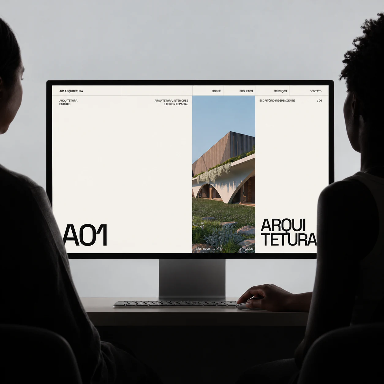 Duas pessoas observam a página inicial da A01 Arquitetura em um monitor.